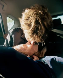 Close-up portrait of two people kissing inside a car with warm natural sunlight illuminating the scene.