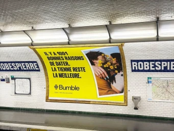 Bumble out of home advertising poster displayed on a white tiled subway station wall featuring a couple holding yellow flowers.