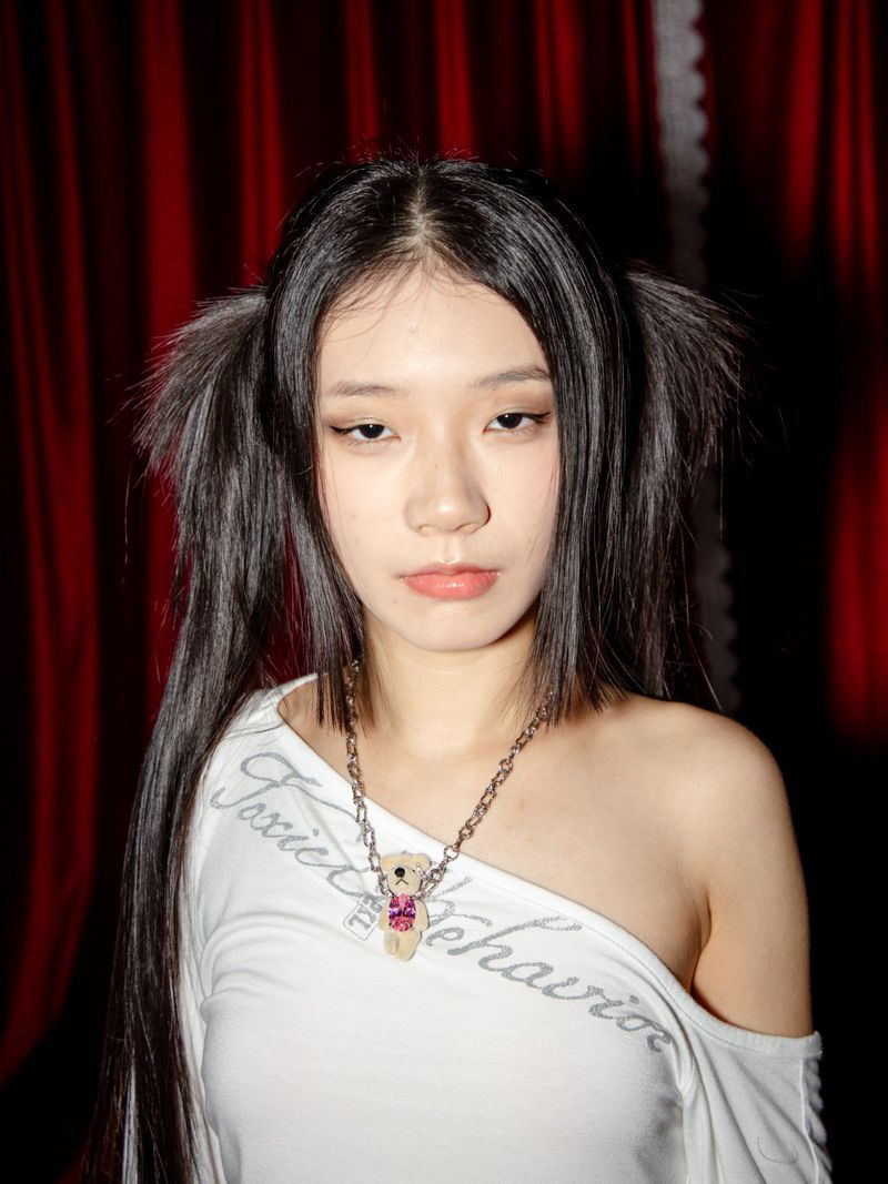 Portrait of an Asian adult female with long black pigtails, wearing a white shirt and bear necklace, standing in an indoor setting with red curtains.