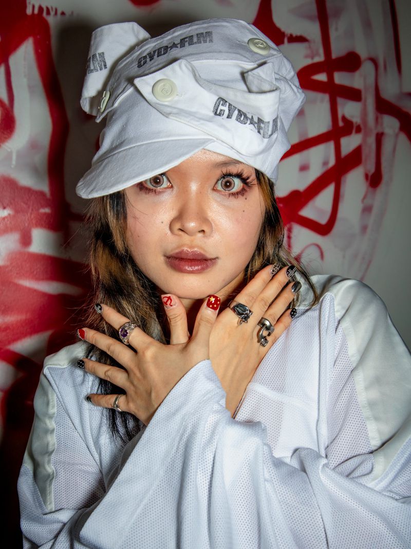 Portrait of a young adult female with light skin wearing a white hat and loose-fitting mesh shirt, set against a graffiti background.