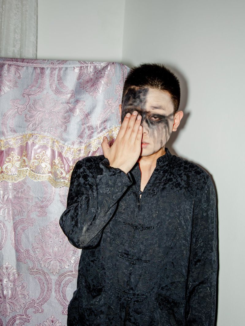 Portrait of an Asian man with black eye makeup, covering his eye with his hand, wearing a black patterned top against a patterned curtain.