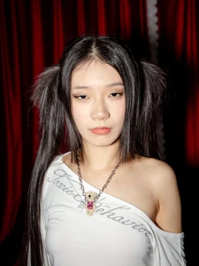 Young East Asian woman with dark pigtails wearing an off-shoulder white top and teddy bear necklace against red velvet curtains.