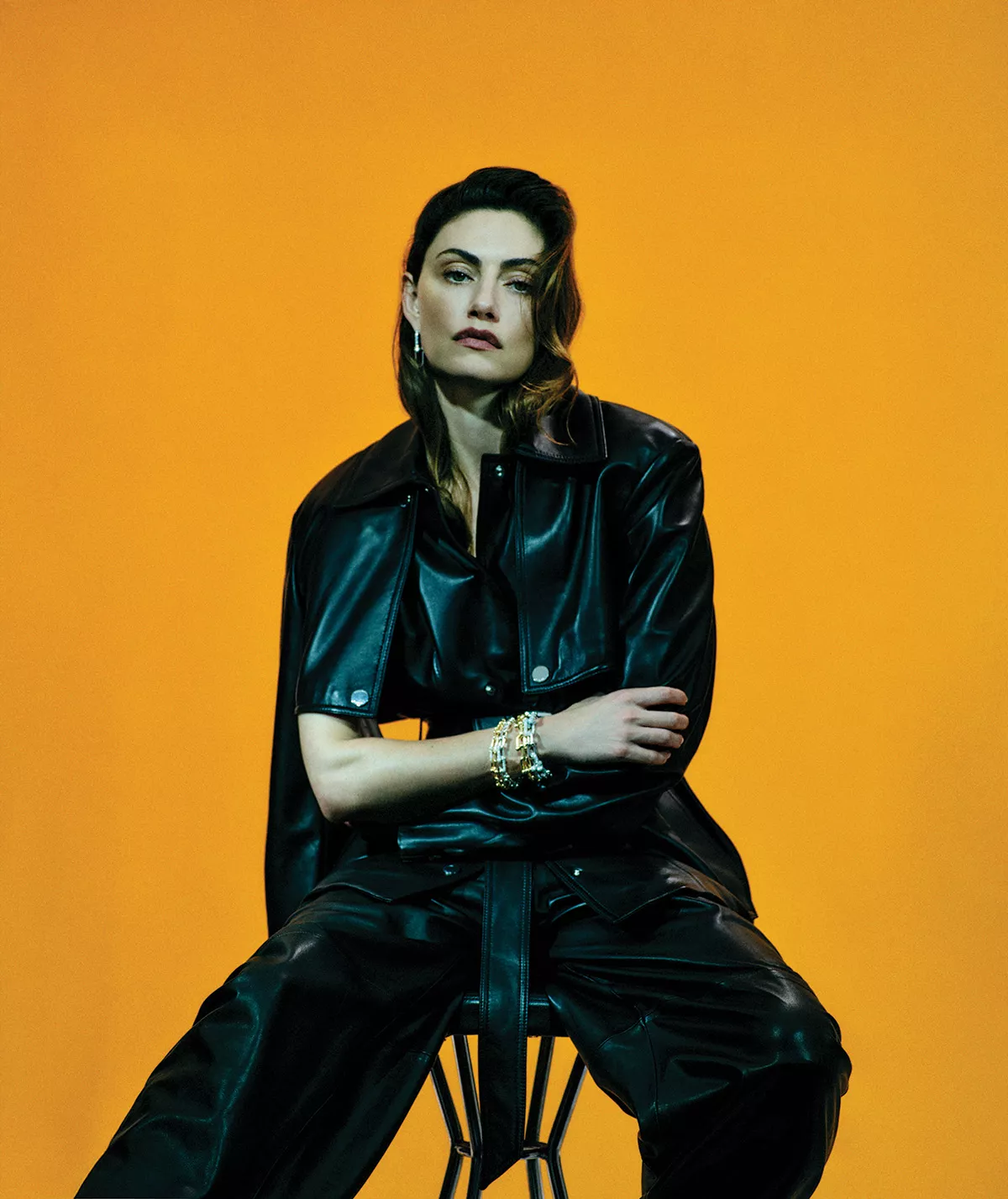 Phoebe Tonkin seated wearing a black leather jacket and trousers against a vibrant orange backdrop