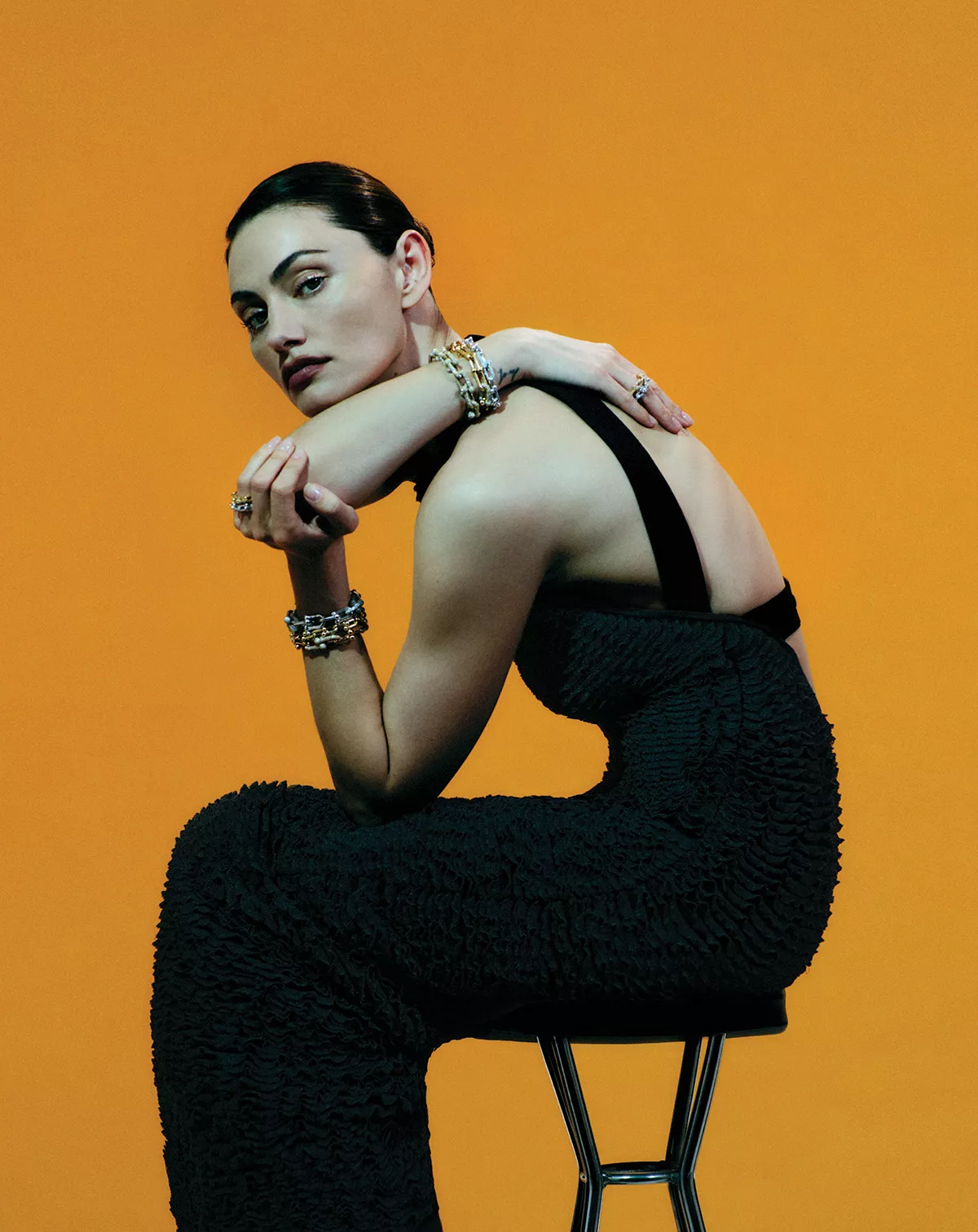 Phoebe Tonkin seated on a stool wearing a textured black dress against an orange background