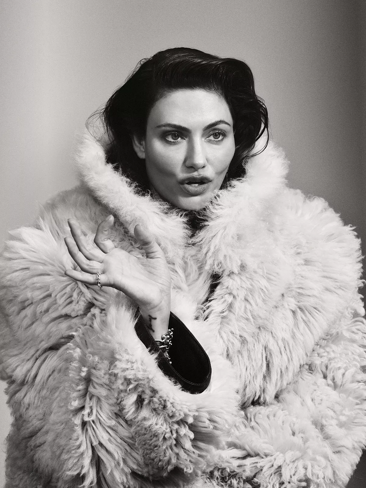 Monochrome portrait of Phoebe Tonkin wearing a fluffy shearling coat against a plain gradient background.