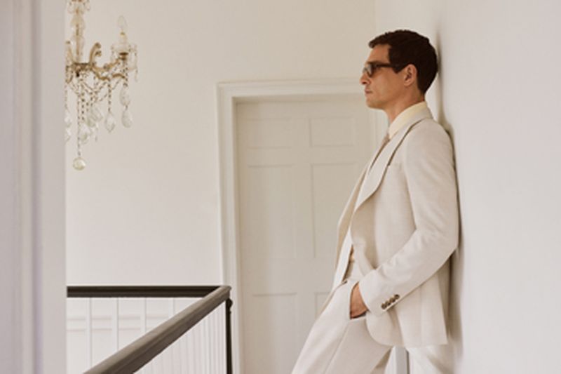Man in a cream suit and glasses leaning against a white wall in an interior hallway with a crystal chandelier.