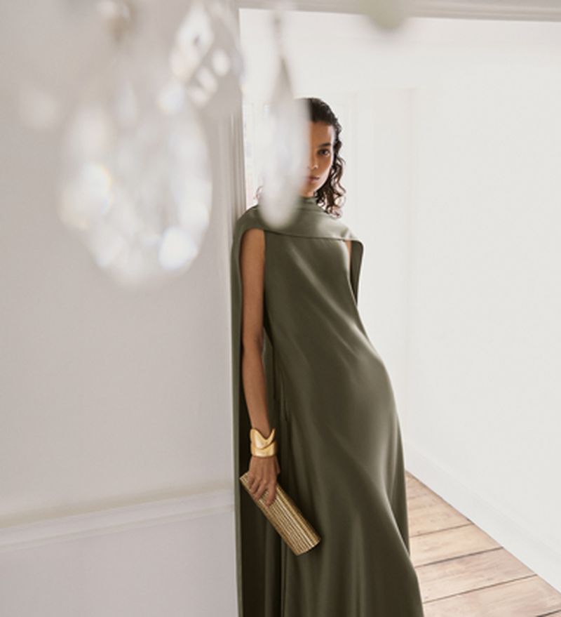 Model wearing an olive green sleeveless dress and gold cuff bracelet holding a clutch inside a bright interior