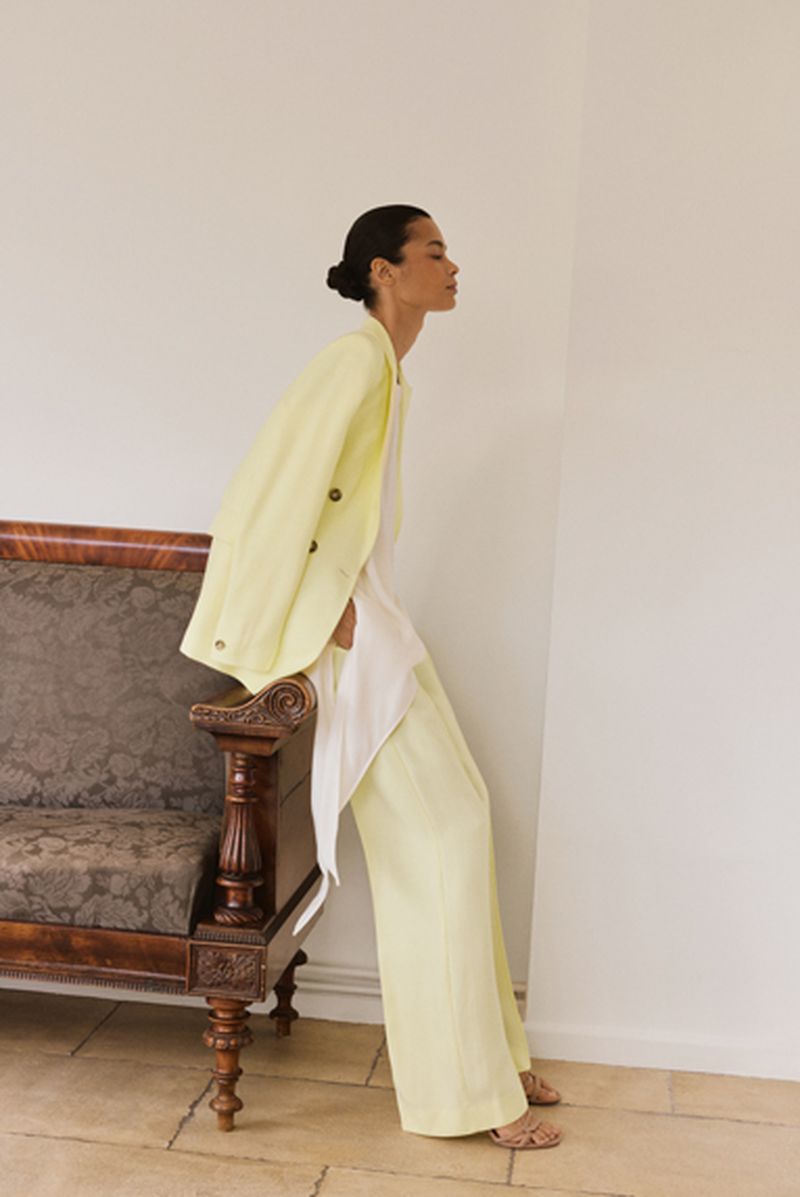 Model wearing a pale yellow blazer and matching trousers leaning against a vintage wooden sofa in an interior setting.