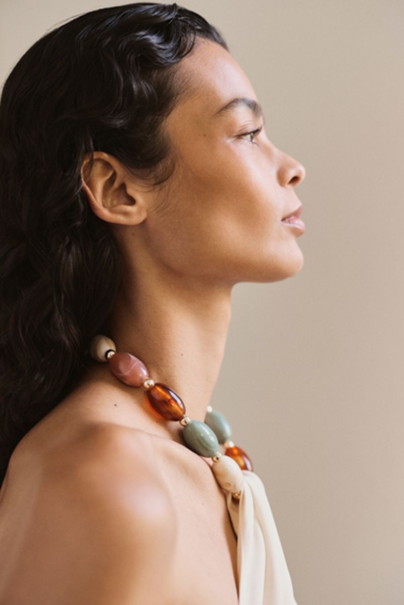 Profile portrait of a woman wearing a beaded statement necklace and tied fabric top against a plain neutral background.