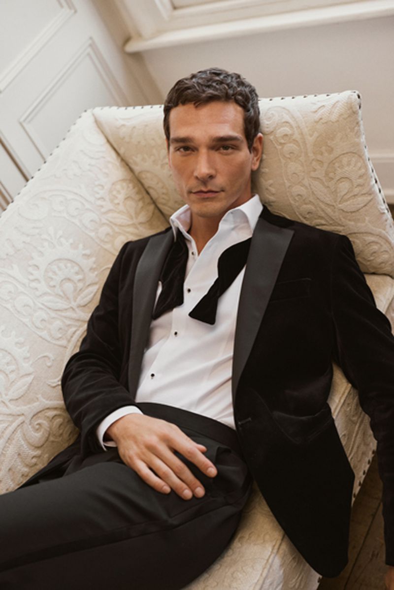 Man seated on an ornate patterned sofa wearing a black velvet tuxedo jacket and an untied bow tie for Reiss.
