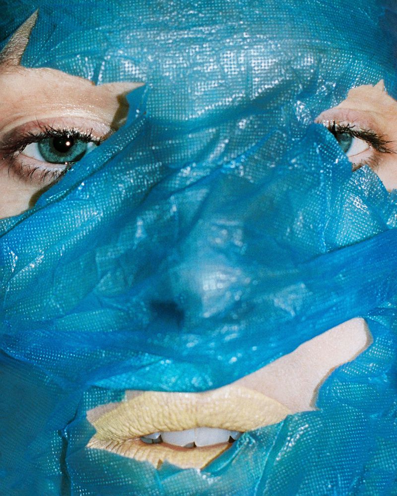 Close-up portrait of a person's face partially covered with a blue plastic material, highlighting their eyes and mouth area.