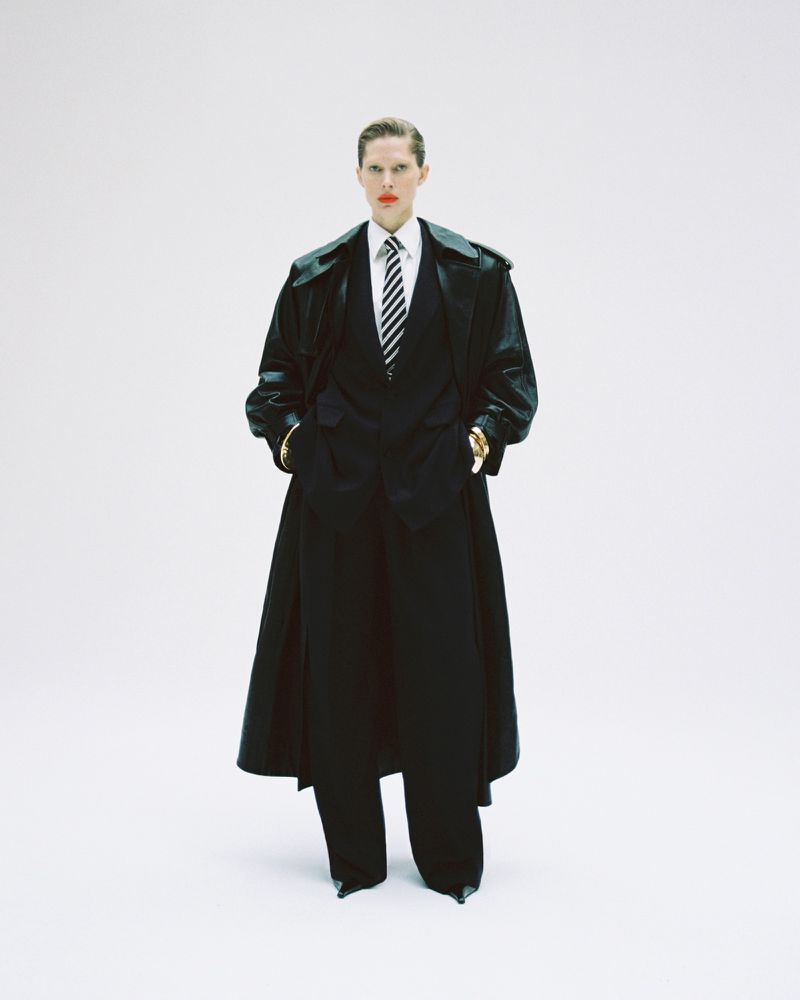 A model in a black coat and suit poses in a stark white setting, accented by a striped tie and gold bracelets, for a fashion editorial.