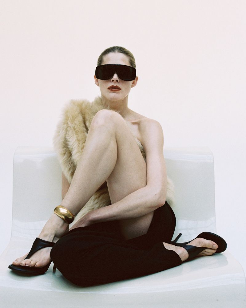 An adult female model with sunglasses and draped in a fur shawl sits on a white chair in a studio environment. She is wearing black heels and trousers.