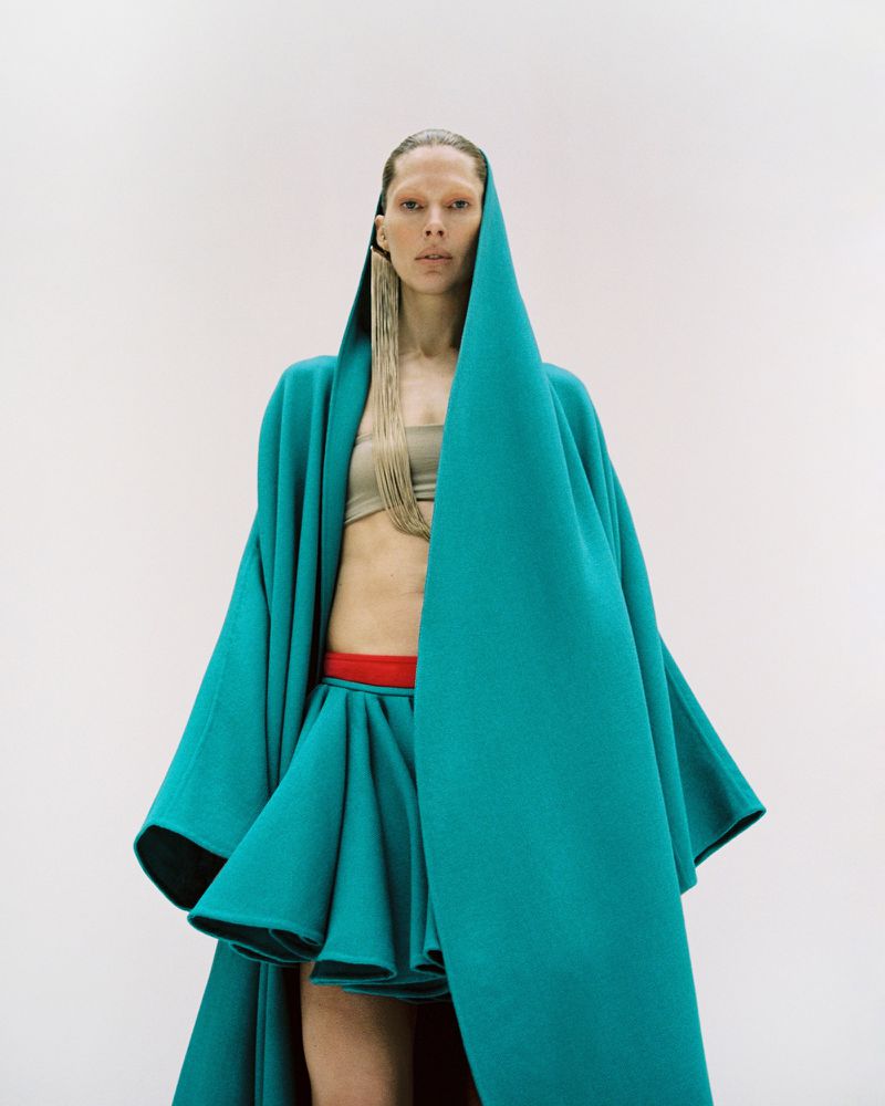 Fashion shot: Woman in teal hooded coat and skirt against a white background, with long blonde tassles hanging from her hair.