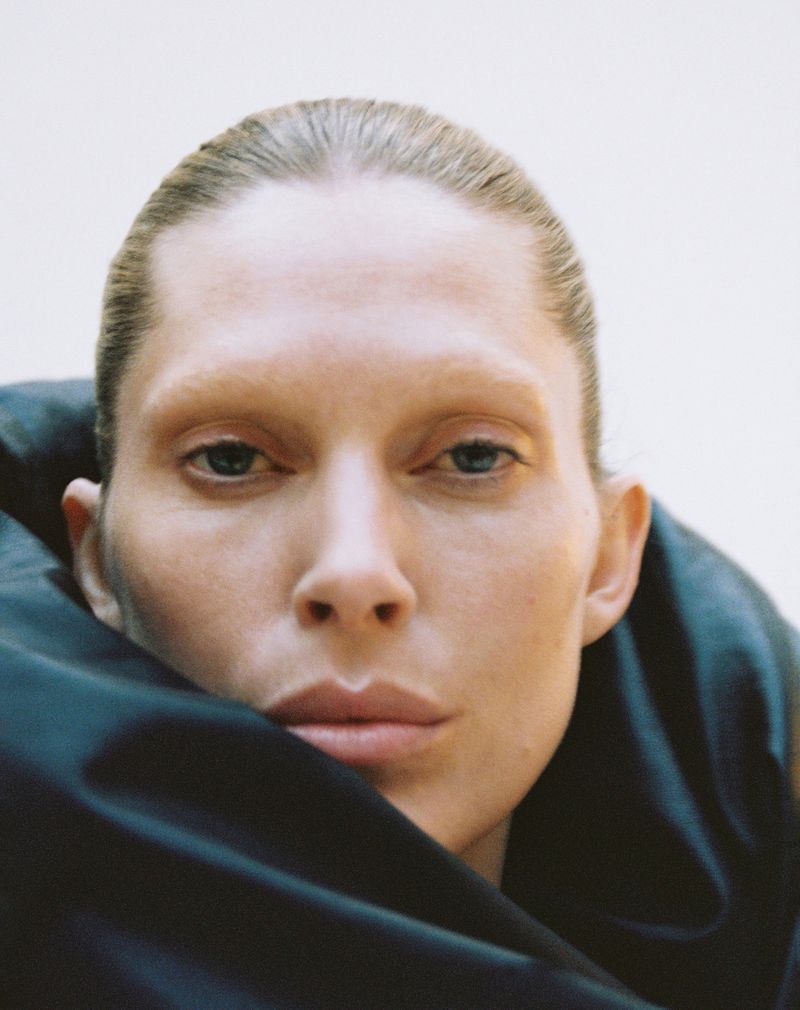 Close-up portrait of a woman with light skin and slicked-back blonde hair, wearing a black draped garment against a neutral background.