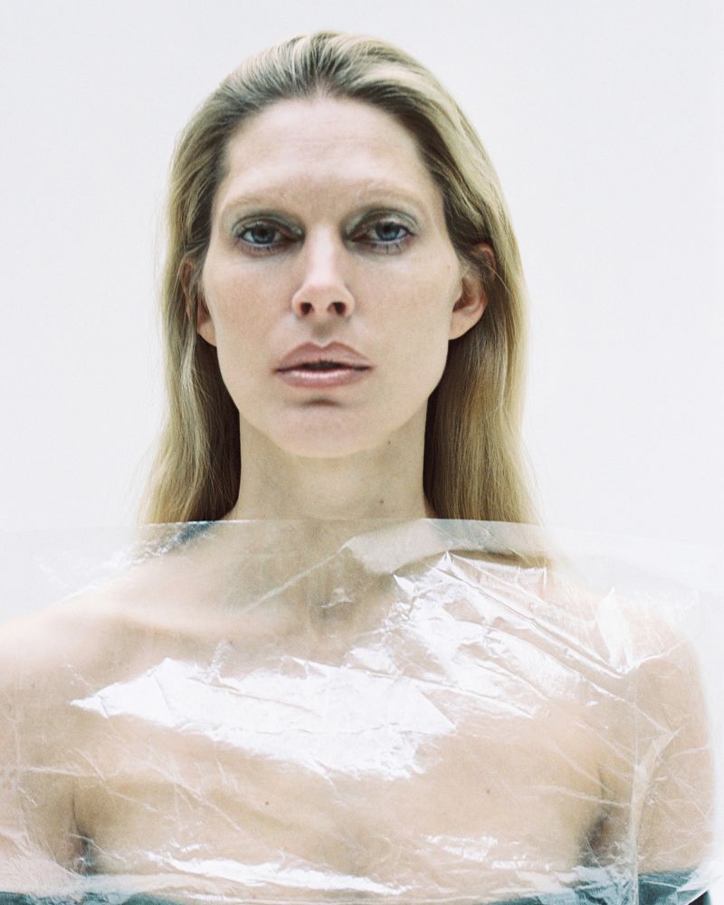Blonde woman in a close-up portrait, wrapped in translucent plastic against a stark white background. Studio lighting.