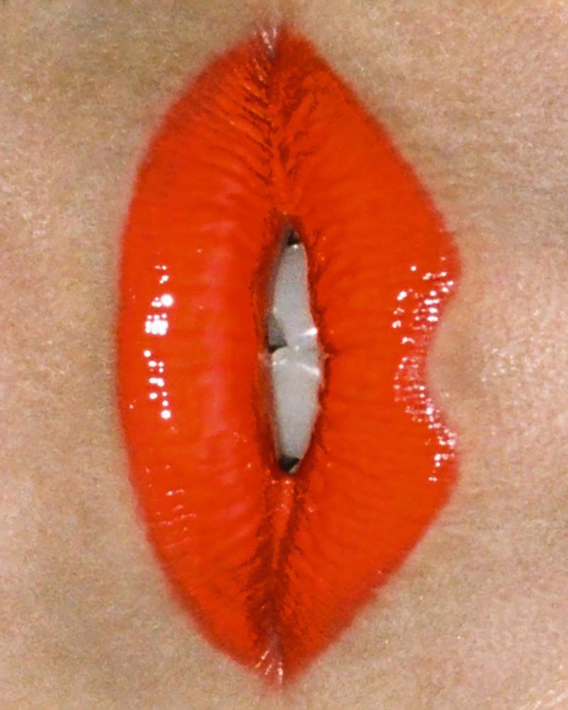 Close-up of a woman's red, glossy lips parted to show teeth. The focus highlights the vibrant color and texture of the lipstick.