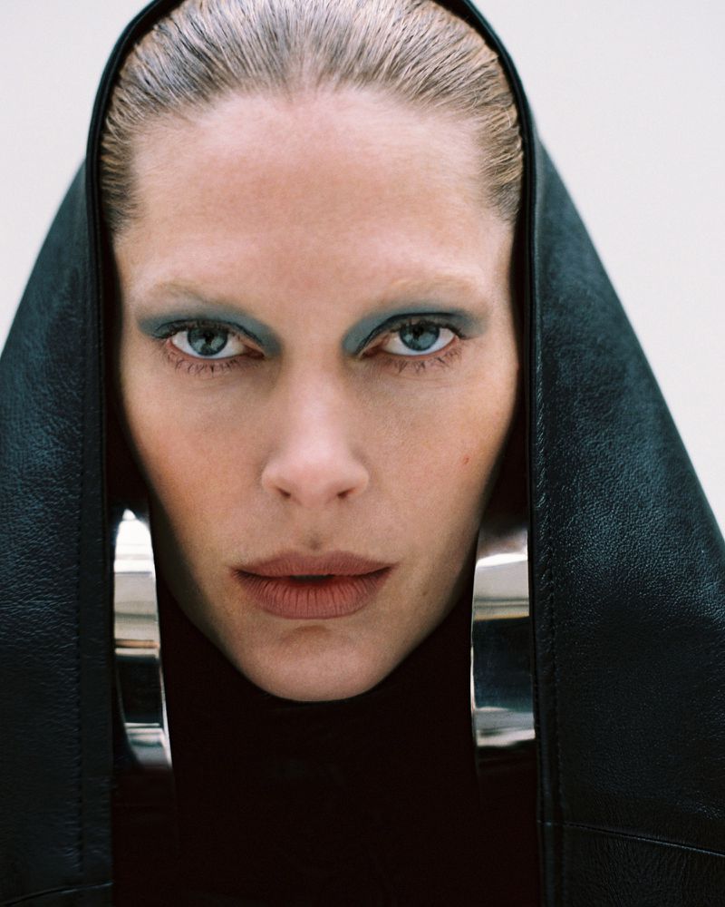 Close-up of a woman in black clothing with large silver hoop earrings and green eyeshadow, set against a white background.
