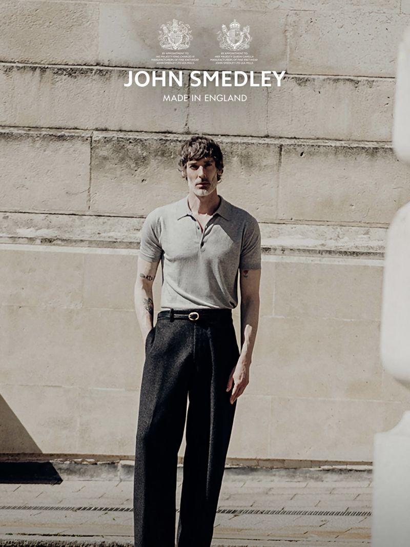 Model Richard Biedul wearing a grey polo shirt and black trousers against a stone building exterior for John Smedley.
