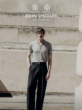 Model Richard Biedul wearing a grey polo shirt and black trousers against a stone building exterior for John Smedley.