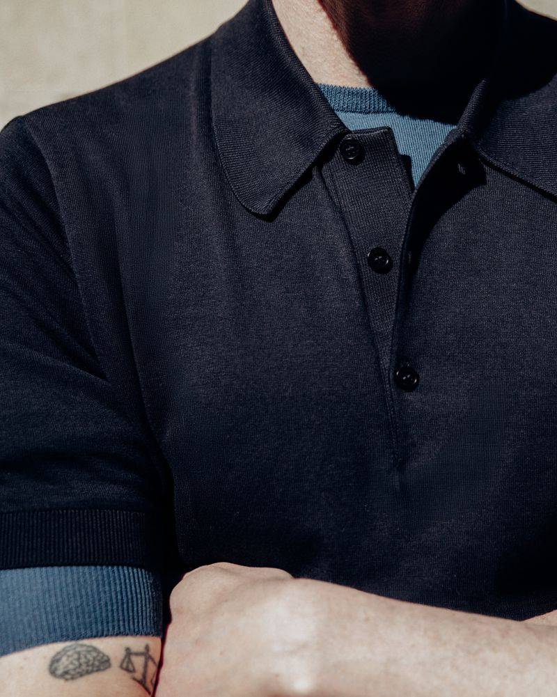 Close-up portrait of Richard Biedul wearing a dark polo shirt with a blue underlayer for John Smedley shot by Jonathan Daniel Pryce