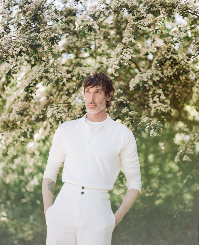 Richard Biedul wearing a white knit polo shirt outdoors against a dense blossoming tree for John Smedley.