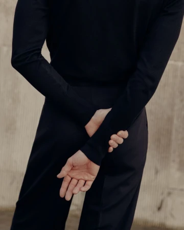 Man viewed from behind wearing a black long-sleeved polo shirt and dark trousers with hands clasped behind his back