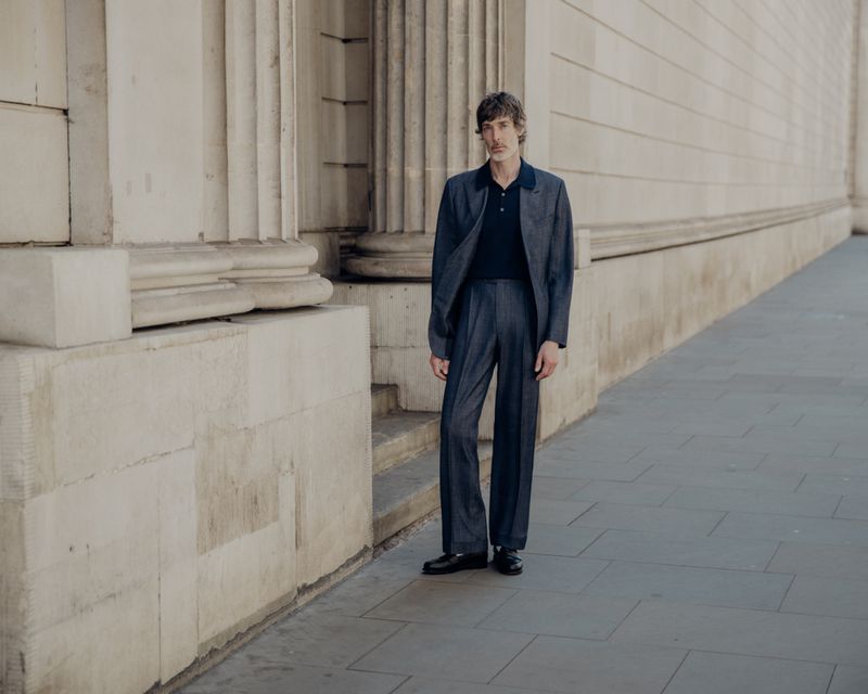 Richard Biedul standing on a paved street beside neoclassical columns wearing a dark grey suit and polo shirt by John Smedley.