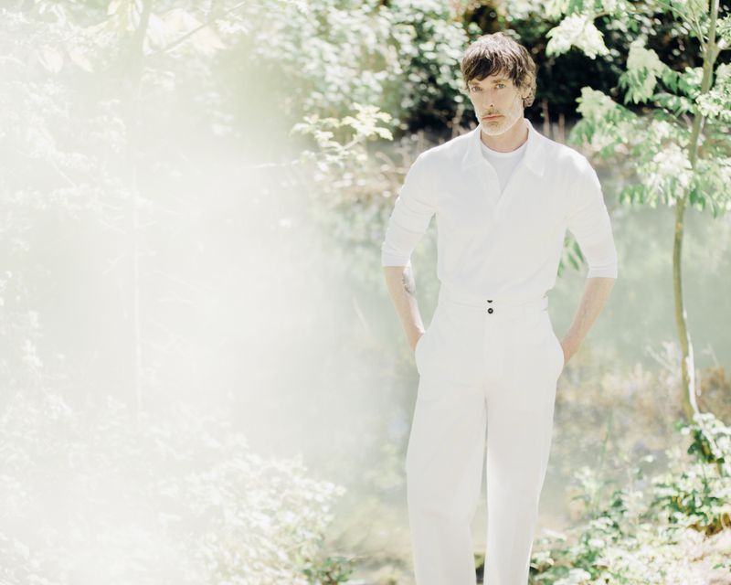 Model Richard Biedul wearing a white polo shirt and white trousers outdoors with soft leafy foliage in the background.