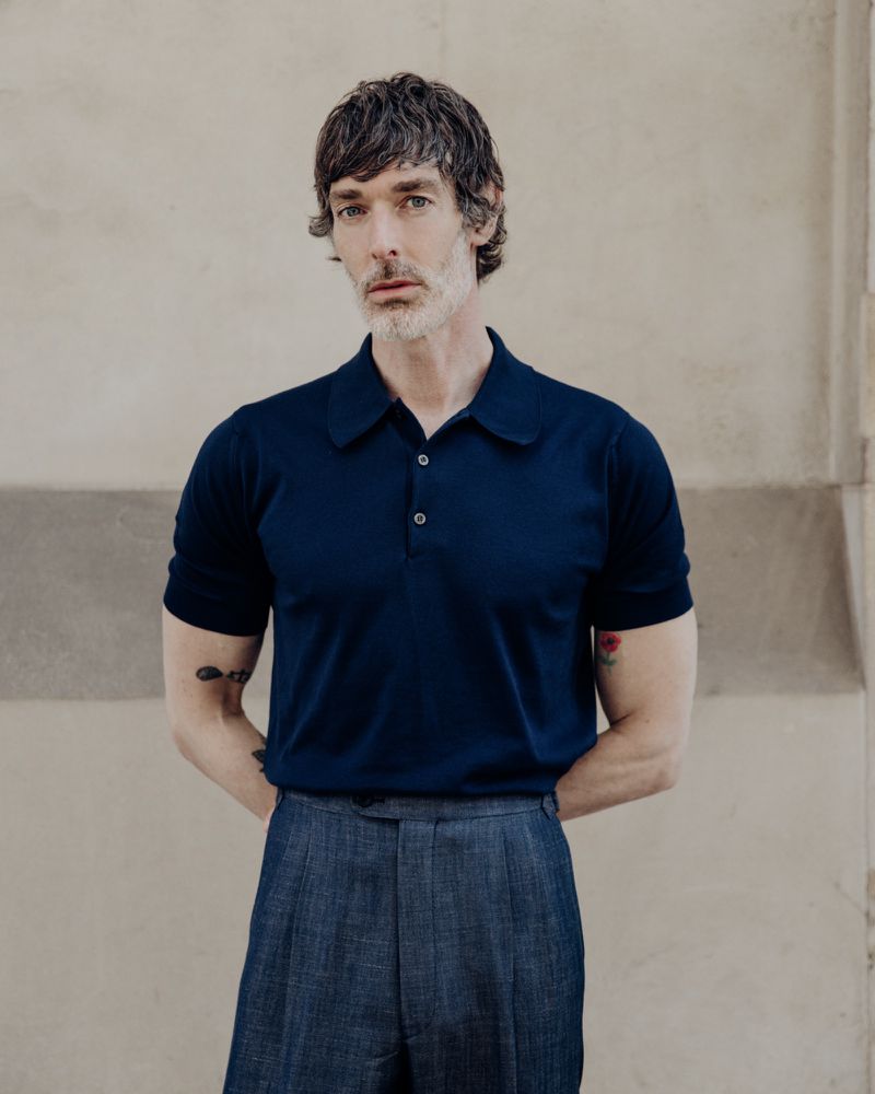 Model Richard Biedul wearing a navy polo shirt and grey trousers against a neutral wall for John Smedley.