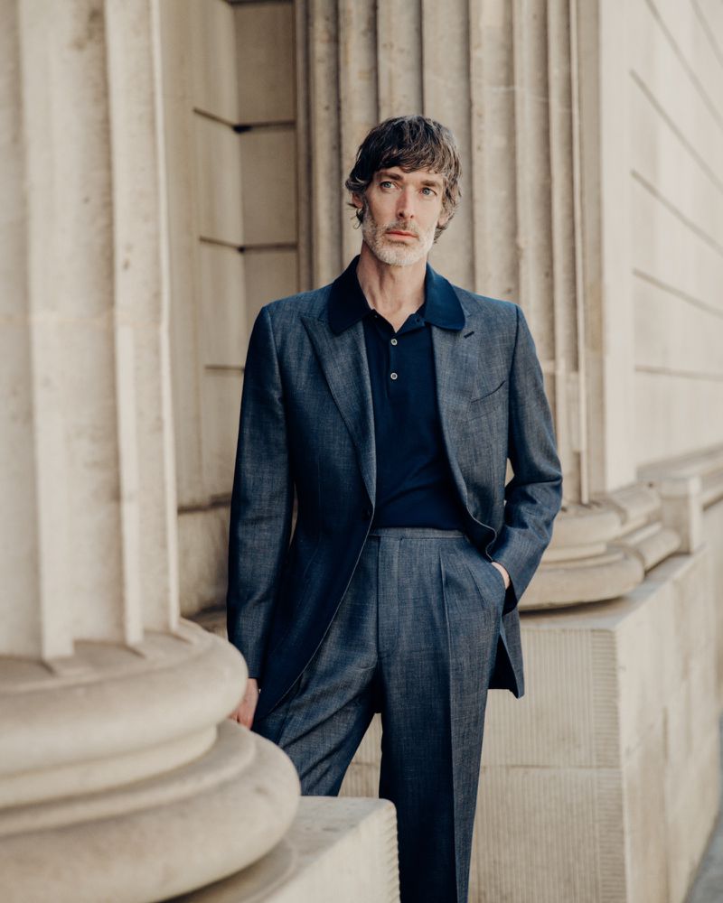 Model Richard Biedul wearing a navy polo shirt and grey suit standing outdoors against classical stone architecture