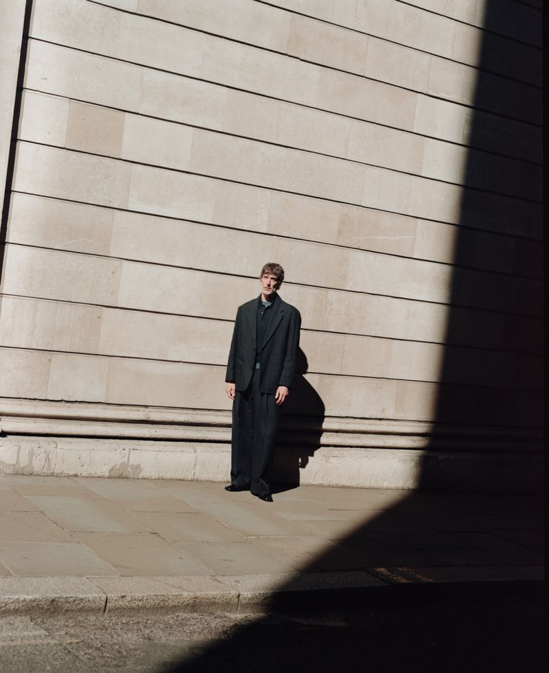 Model standing outdoors against a large stone ashlar wall under strong directional sunlight and shadow for John Smedley.