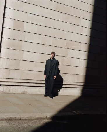 Model standing outdoors against a large stone ashlar wall under strong directional sunlight and shadow for John Smedley.