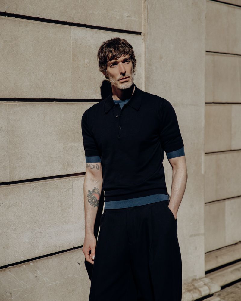 An adult male poses wearing a navy polo shirt and dark pants against a textured wall, with natural light enhancing his features.
