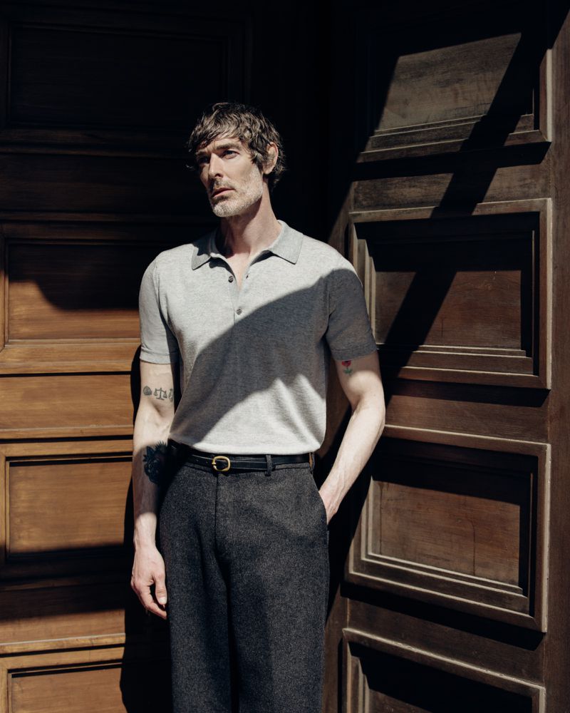 Man wearing a grey polo shirt and dark trousers standing in front of a dark wooden panelled door.