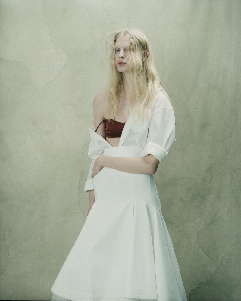 A fair-skinned young woman with blonde hair poses wearing a white dress and a partially open white shirt over a maroon top against a textured background.