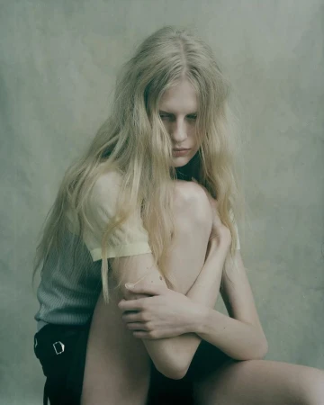 Blonde woman with long wavy hair sits with arms wrapped around legs in a minimalist studio portrait against a mottled light green background.