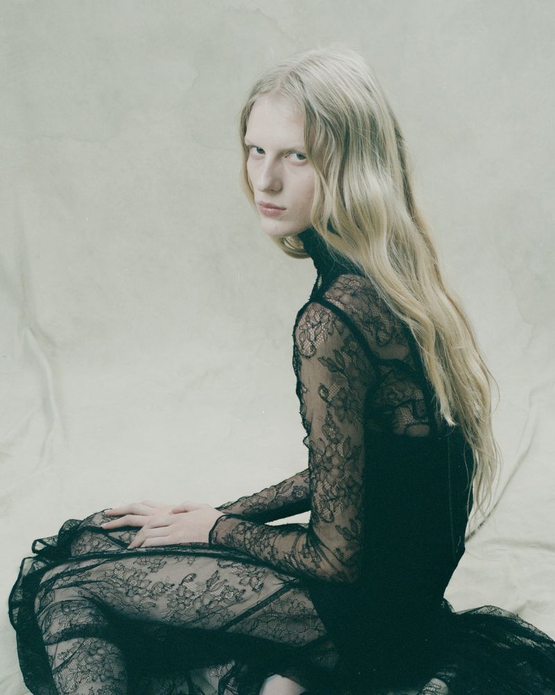 Woman with long wavy blonde hair poses while sitting down, wearing a long-sleeved lace dress, in a photo studio with a seamless backdrop.