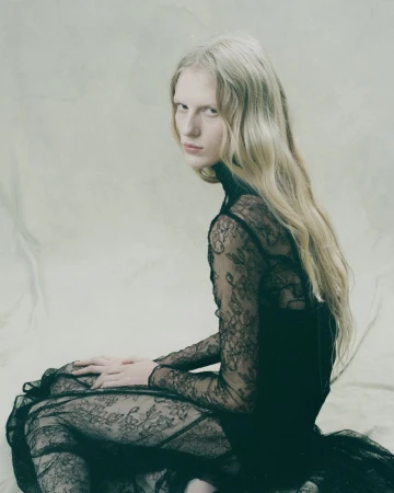 Woman with long wavy blonde hair poses while sitting down, wearing a long-sleeved lace dress, in a photo studio with a seamless backdrop.