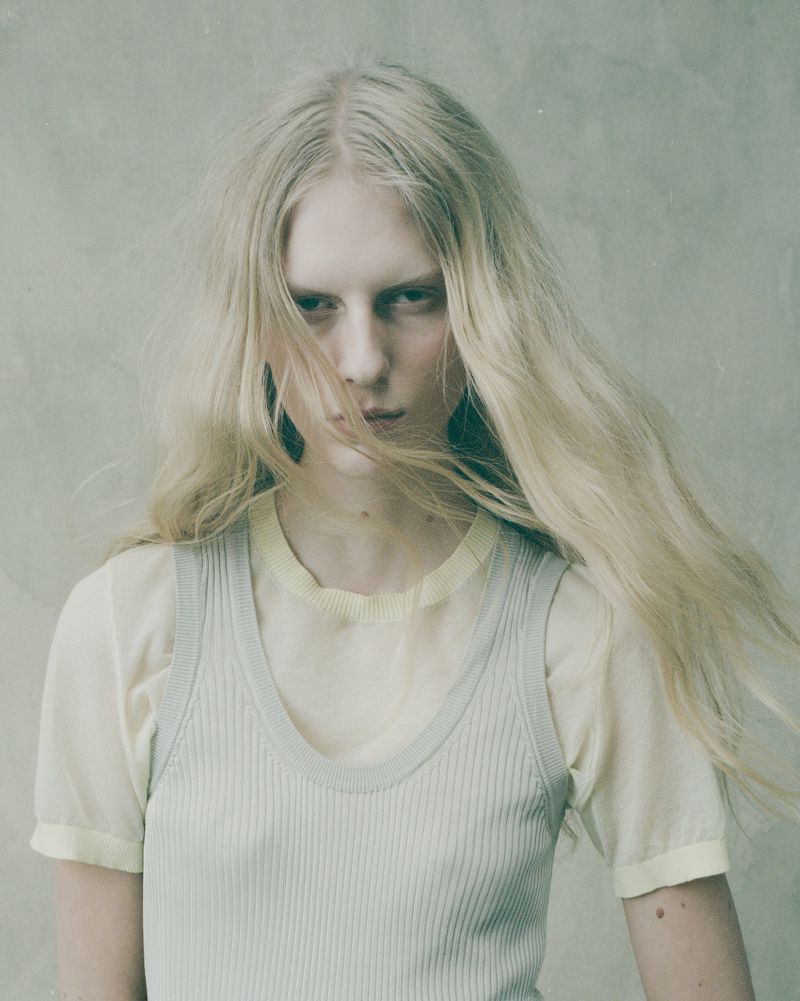 Close-up portrait of a young adult woman with long blonde hair, wearing a light blue top and a pale yellow shirt, posing indoors.