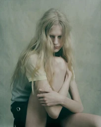 Model with long blonde hair wearing a light top and dark skirt seated with knees pulled up against a neutral textured studio backdrop.