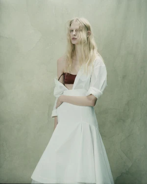 Model with long blonde hair wearing a white shirt and skirt with a red bandeau against a textured green background.