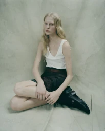 Model Eduarda Muehlmann seated against a neutral textured backdrop wearing a white tank top and dark skirt