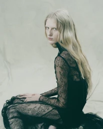 Model Eduarda Muehlmann wearing a sheer black floral lace dress seated against a textured pale studio backdrop.