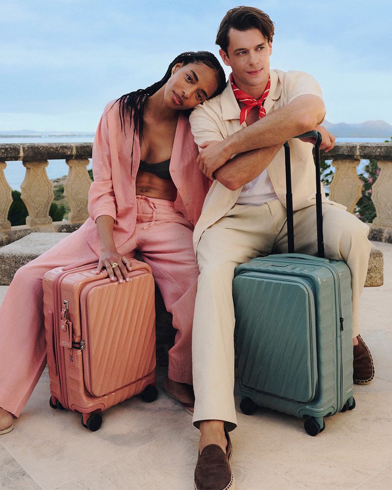 Two models sitting outdoors against a stone balustrade with luggage, featuring a pink suit and a green suitcase for Tumi
