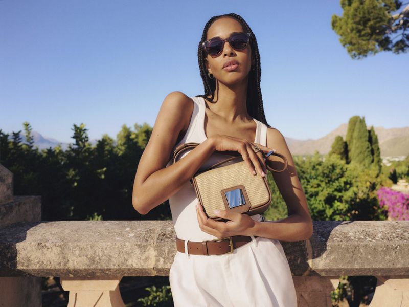 Model wearing sunglasses and white top holding a woven crossbody handbag outdoors against a scenic landscape backdrop