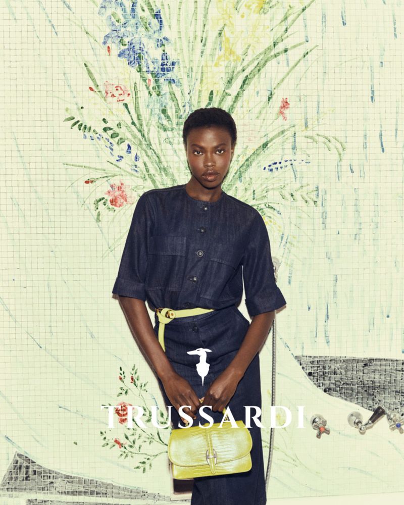 Fashion shot of an African-American woman in a dark denim dress with a yellow belt and purse, standing in front of a floral mural.