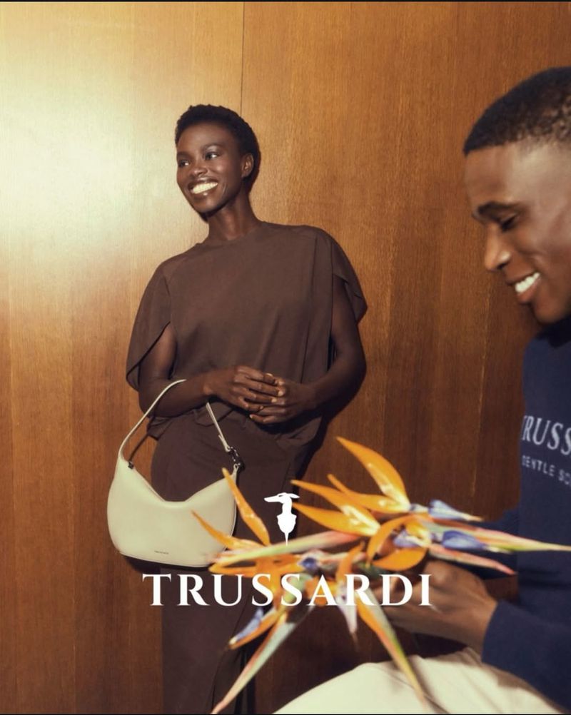 Black woman in brown dress holding cream handbag standing next to man holding flowers for Trussardi SS26 campaign