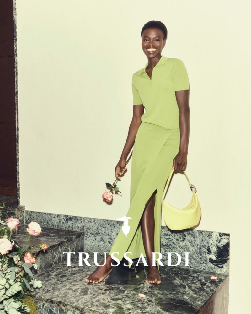 Model smiling on marble steps holding a rose and a yellow handbag in a lime green knit top and skirt set for Trussardi SS26.