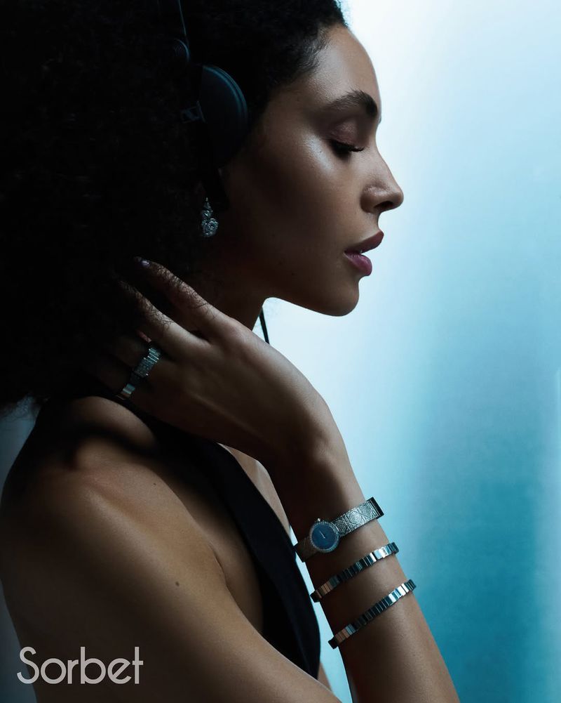 Profile portrait of a woman with dark hair wearing headphones, jewelry, and a black top against a light blue studio backdrop.